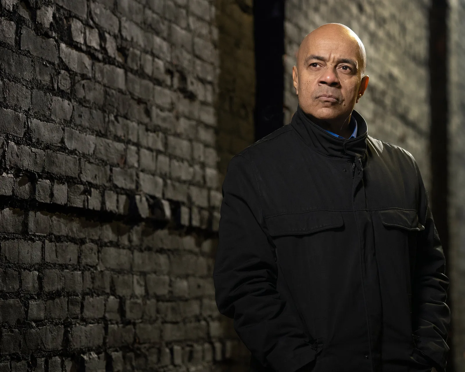 Dramatic editorial portrait of a man against a dark brick wall, photographed by Len Marks